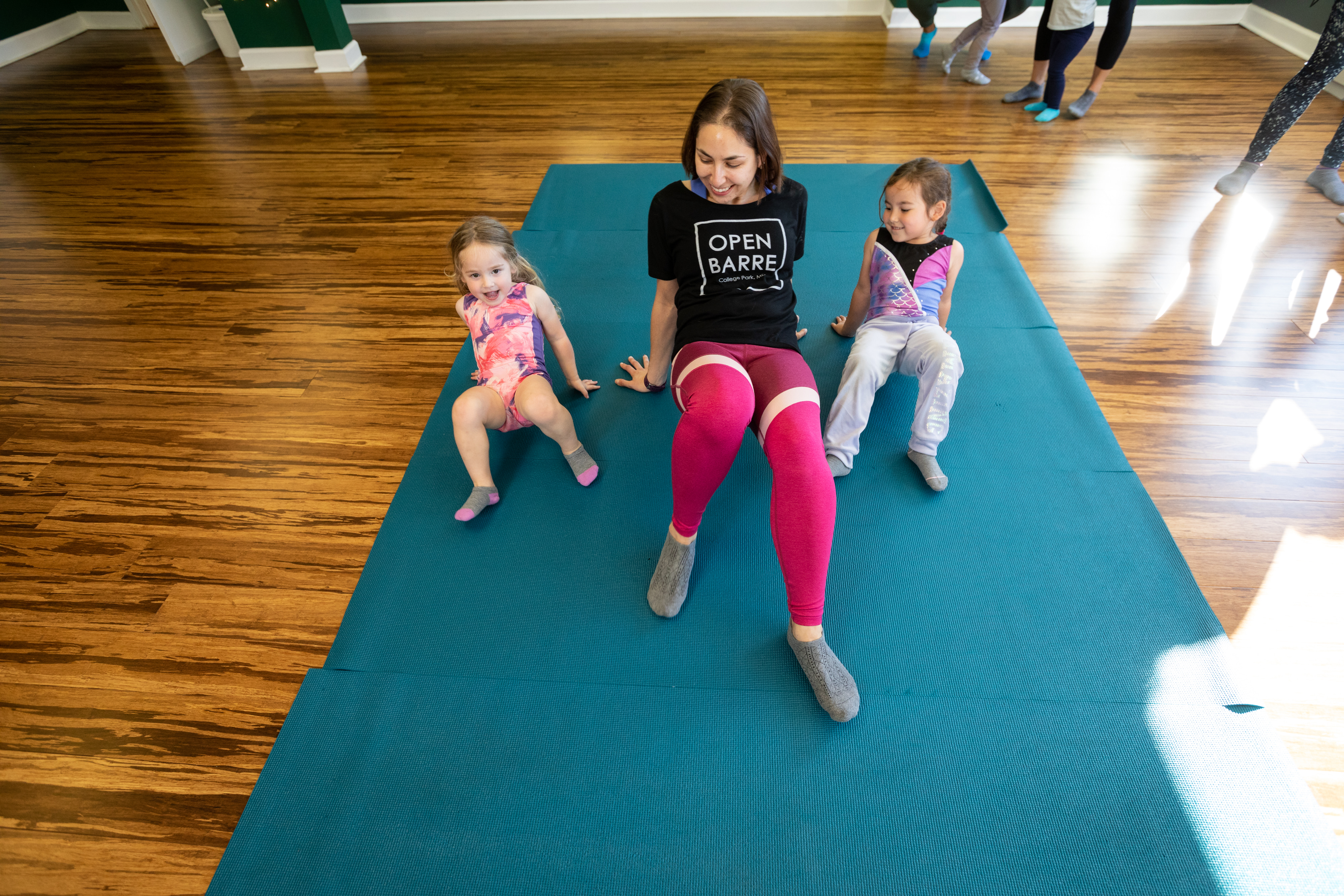 young mom with two daughters in gymnastics leotards walking like crabs on yoga mats in openbarre parent and me class